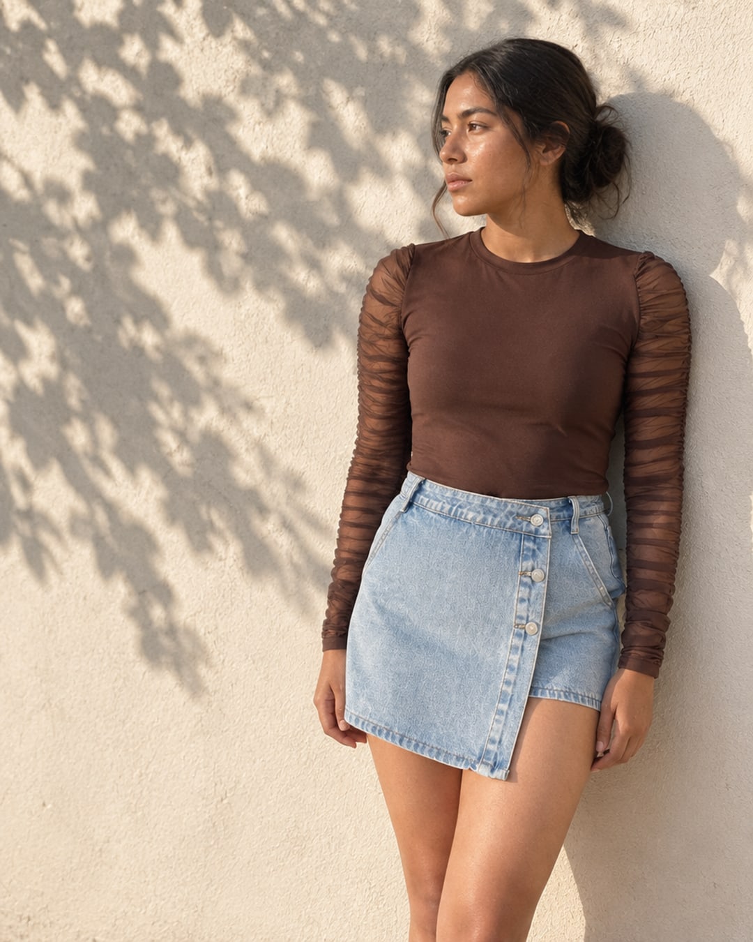 Campaña: el top marrón de manga de malla con la falda-short vaquera, apoyada en un muro.
